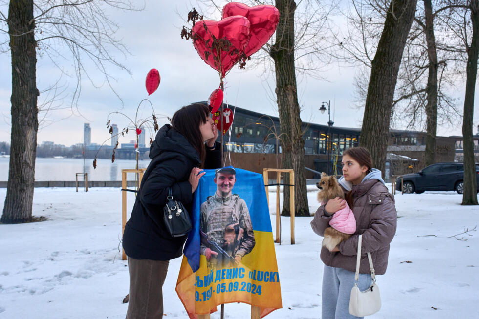 A widow of a fallen soldier, his portrait on the foreground, kisses his ID tag standing with her daughter under balloons in the shape of red hearts on a sakura tree on Valentine's Day on the Love Alle